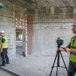 Two workers in hard hats and hi-vis jackets use a laser level in a partially built concrete space